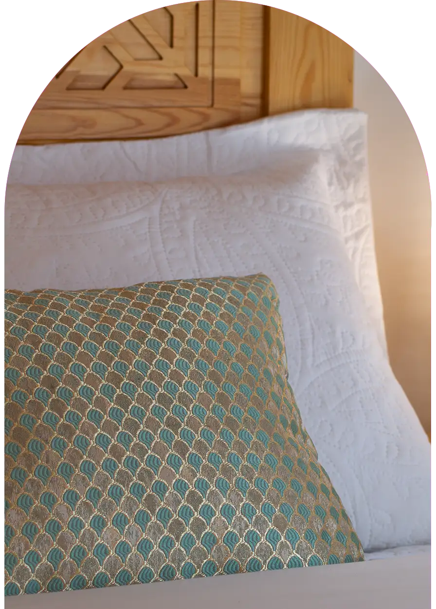 Gros plan sur un oreiller en tissu satiné avec broderies dorées et tête de lit sculptée, détails de décoration d’un riad à Essaouira